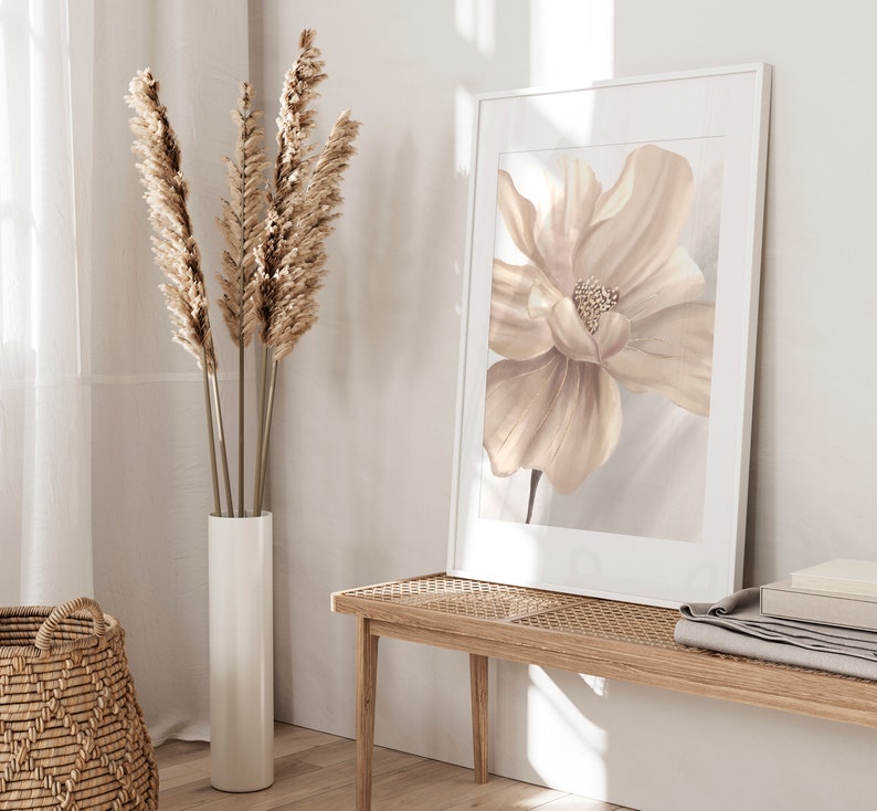 May include: A beige and white floral print in a white frame hangs on a white wall above a light wood bench. A vase with dried pampas grass sits on the floor next to the bench.