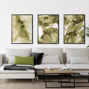 May include: Three framed abstract art prints with green and gold swirling patterns. The prints are hung on a white wall above a white sectional sofa with green and black throw pillows.