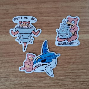 May include: Three cartoon stickers on a wooden surface. One sticker features a pink creature with the text "FIGHT ME BRO". Another shows a pink creature with the text "UNDERTHINKER". The third sticker depicts a pink creature with a blue and white shark.