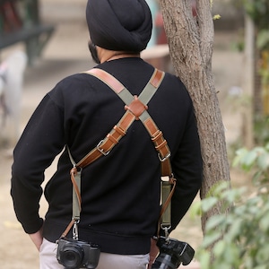 May include: A person wearing a black shirt and a black turban with a brown and green camera strap. Two cameras are attached to the strap, one on each side.