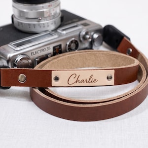 May include: A brown leather camera strap with the name "Charlie" engraved on a light tan leather patch. The strap is coiled next to a vintage silver camera.