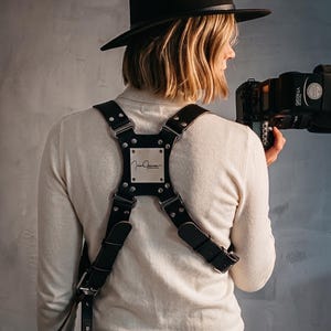Puede incluir: Un arnés de cuero negro para cámara con correas ajustables, herrajes de metal plateado y una placa cuadrada central con el texto grabado "Jrie James". El arnés se lleva sobre una camisa de manga larga color crema y un sombrero negro.