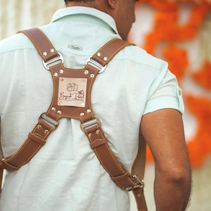 May include: A brown leather camera strap with adjustable straps and a metal buckle. The strap has a rectangular patch with the text "Byresh Patel Photography" in a brown font.