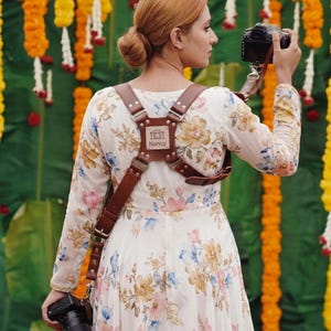 Puede incluir: Un arnés de cámara de cuero marrón con el nombre "Nancy" y un icono de cámara. El arnés se usa sobre un vestido floral blanco. Una cámara se sostiene en cada mano. El fondo presenta decoraciones florales amarillas y blancas.