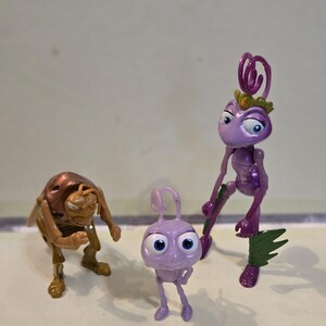Puede incluir: Tres figuras de plástico de personajes de Bichos. A la izquierda, un saltamontes marrón en postura encorvada. En el centro, una pequeña hormiga de color violeta claro con grandes ojos azules. A la derecha, una hormiga alta violeta con corona dorada.