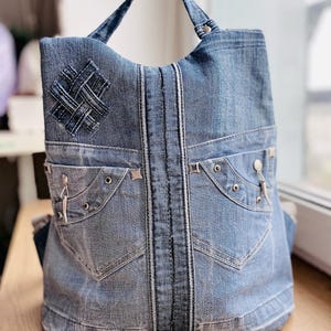 May include: A denim tote bag made from recycled jeans. The bag features the pockets and seams of jeans, with a handle and a decorative patch. The bag is light blue and sits on a wooden surface. The bag is a unique design.