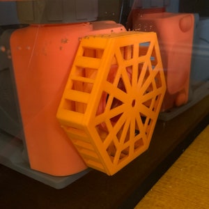 Puede incluir: Un filtro naranja geométrico en forma de panal se encuentra en un acuario transparente. El filtro tiene un diseño circular en el centro. Detrás del filtro hay componentes naranjas y grises. Un objeto cilíndrico blanco descansa sobre una superficie de madera.