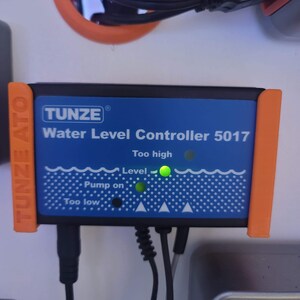 May include: A blue Tunze Water Level Controller 5017 with orange accents. The device has a series of indicator lights, including a green "Level" light. Black cables are connected to the device.