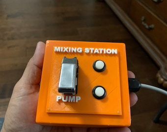 May include: An orange mixing station with the text "MIXING STATION" and "PUMP" in white. It features a silver switch and two white buttons. A grey cable extends from the side. The device is held in a hand.