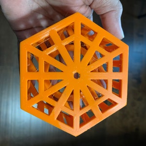 May include: An orange, geometric, hexagonal object with an open, lattice-like design. The object has a central hole and is held in a hand. The design features a series of interconnected triangles and lines, creating an openwork pattern.