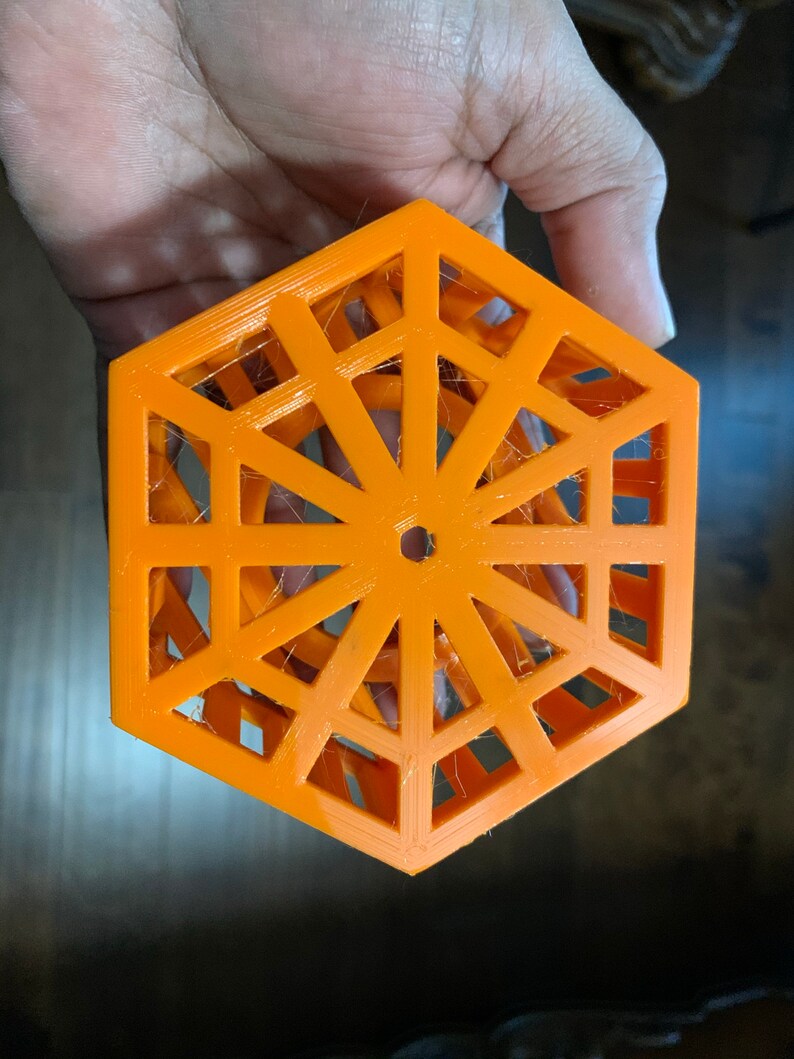 May include: An orange, geometric, hexagonal object with an open, lattice-like design. The object has a central hole and is held in a hand. The design features a series of interconnected triangles and lines, creating an openwork pattern.