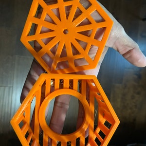 May include: Two-piece orange hexagonal object. The top piece has a geometric design with a central hole. The bottom piece is a cage-like structure with a circular opening. Both pieces are held in a hand.