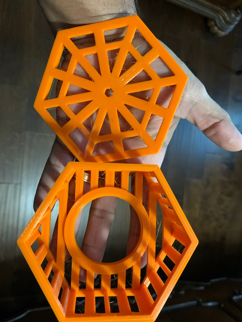 May include: Two-piece orange hexagonal object. The top piece has a geometric design with a central hole. The bottom piece is a cage-like structure with a circular opening. Both pieces are held in a hand.
