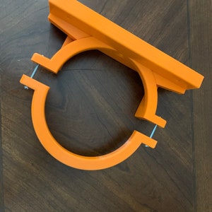 May include: An orange, 3D-printed bracket with a circular base and a rectangular top. Two metal screws secure the circular base. The bracket is designed to hold an object.