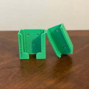 May include: Two bright green 3D-printed InkBird Controller holders. One is upright, and the other is tilted to the side. Each has three holes and a curved bottom. The text "InkBird Controller" is printed vertically on one holder.