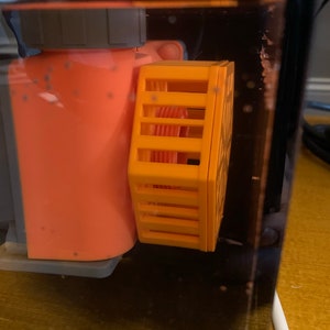 May include: An orange aquarium pump with a yellow protective cage attached. The pump is partially submerged in water, with a clear plastic tank visible. The cage has a rectangular shape with vertical bars.