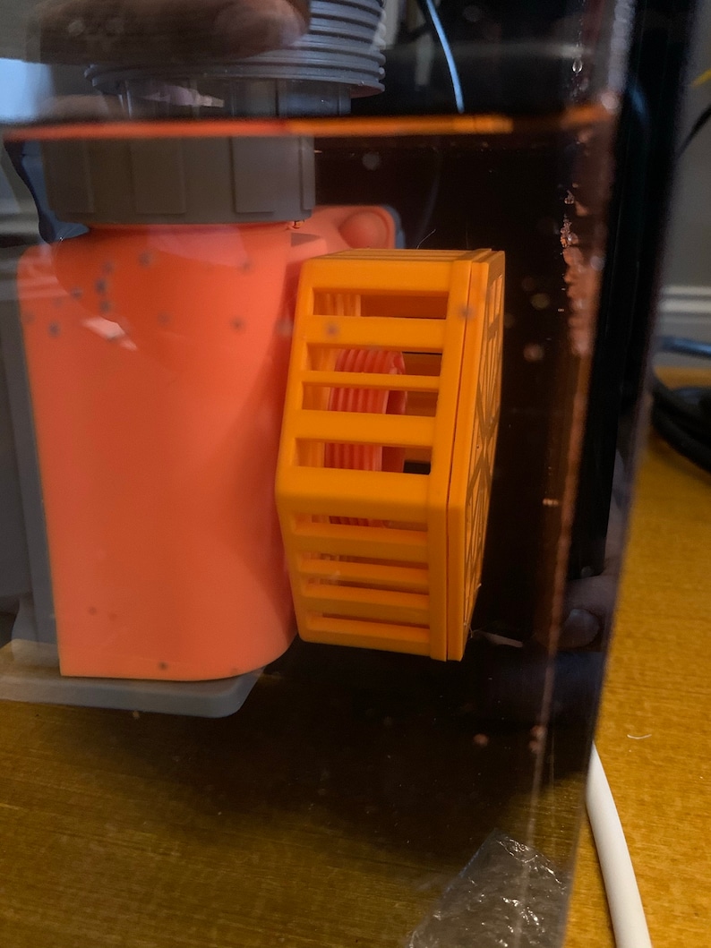 May include: An orange aquarium pump with a yellow protective cage attached. The pump is partially submerged in water, with a clear plastic tank visible. The cage has a rectangular shape with vertical bars.