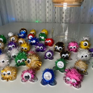 Könnte beinhalten: Eine Sammlung bunter, flauschiger Pompon-Kreaturen mit Glitz Augen. Jeder Pompon ist mit glitzernden, schillernden Fasern verziert und hat eine flache Basis.