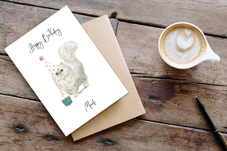 Puede incluir: Una tarjeta de cumplea&ntilde;os blanca con un gato blanco que lleva un gorro de fiesta de lunares rosa y blanco y sostiene un cupcake con una vela encendida. La tarjeta dice "Feliz cumplea&ntilde;os" y "Mark".