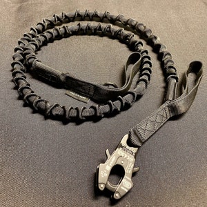 May include: Black tactical dog lead with a bungee cord design. Features a heavy-duty metal clip and a handle. The lead is made of durable nylon and is designed for strength and flexibility. Ideal for medium to large dogs.