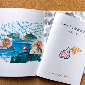 May include: A sketchbook with a blue and white watercolor illustration of a sailboat on a body of water with islands. The cover of the sketchbook is white with the text "SKETCHBOOK VOL. 1" and a watercolor illustration of a fig with a leaf in orange and pink.