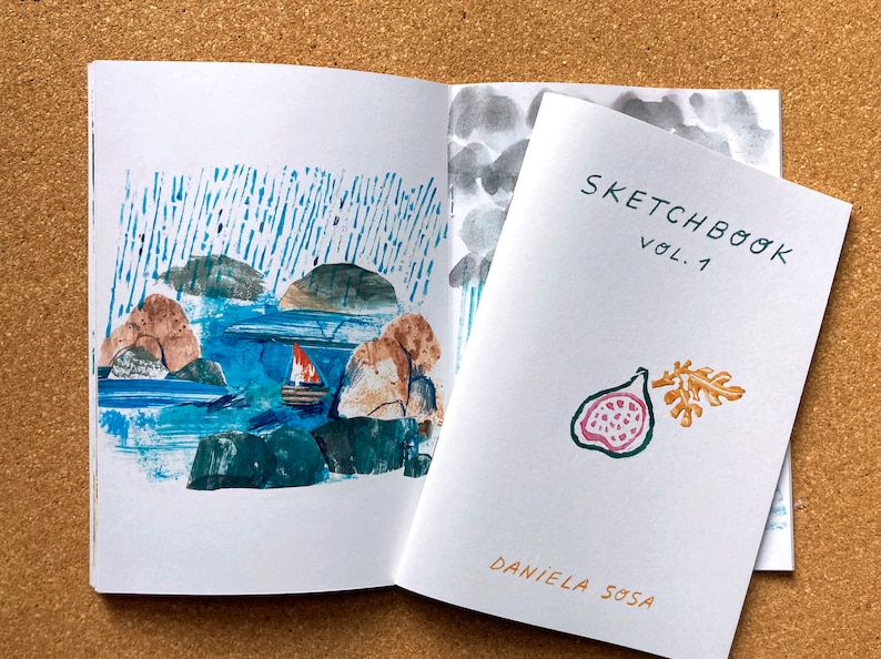 May include: A sketchbook with a blue and white watercolor illustration of a sailboat on a body of water with islands. The cover of the sketchbook is white with the text "SKETCHBOOK VOL. 1" and a watercolor illustration of a fig with a leaf in orange and pink.