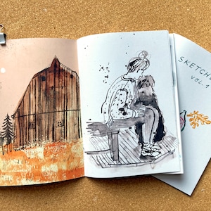 May include: A sketchbook with a black and white illustration of a person sitting on a bench with a dog. The cover of the sketchbook says "SKETCHBOOK VOL. 1" and has a small illustration of a leaf.