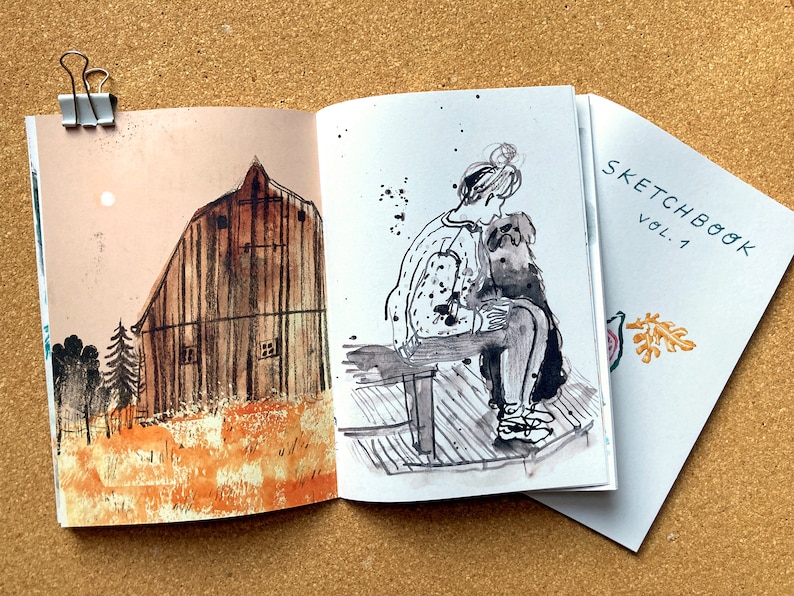 May include: A sketchbook with a black and white illustration of a person sitting on a bench with a dog. The cover of the sketchbook says "SKETCHBOOK VOL. 1" and has a small illustration of a leaf.