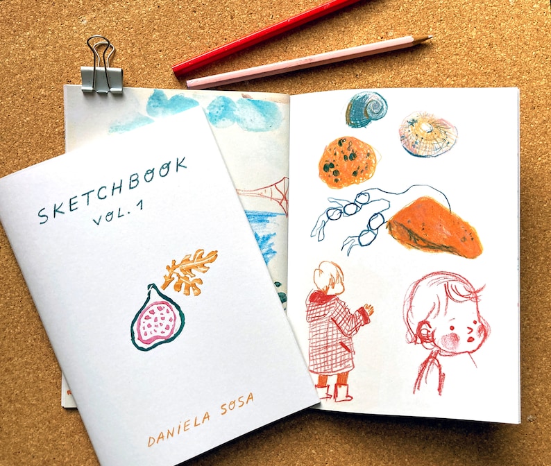 May include: A white sketchbook with the title "Sketchbook Vol. 1" and the name "Daniela Sosa" printed on the cover. The sketchbook is open to a page with hand-drawn illustrations of a fig, a seashell, a crab, a person, and a bridge.
