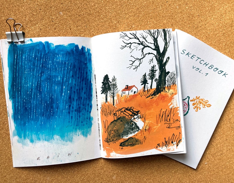 May include: An open sketchbook with a blue watercolor painting of rain on the left page and a watercolor painting of a house in a forest on the right page. The cover of the sketchbook is white with the text "SKETCHBOOK VOL. 1" in black.