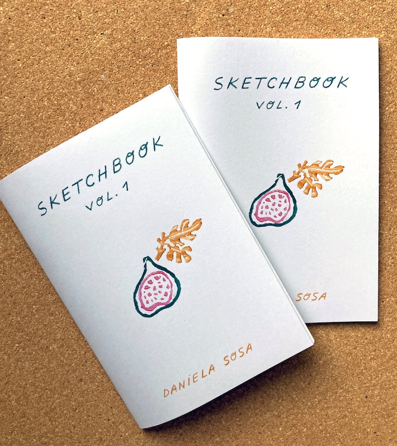 May include: Two white sketchbooks with the title "Sketchbook Vol. 1" printed on the front cover. The covers feature a hand-drawn illustration of a fig with a leaf in gold and brown tones. The bottom of the cover reads "Daniela Sosa".