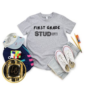 May include: A gray t-shirt with the text "FIRST GRADE STUD(ent)" in black. The shirt is laid out on a white background with a backpack, a hat, and a pair of white sneakers.