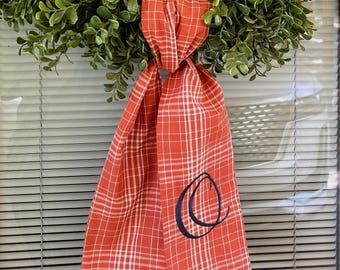 May include: A green, leafy wreath with a red and white plaid ribbon. The ribbon is tied in a knot and features a navy blue embroidered letter "O". The wreath is hanging on a blue door.