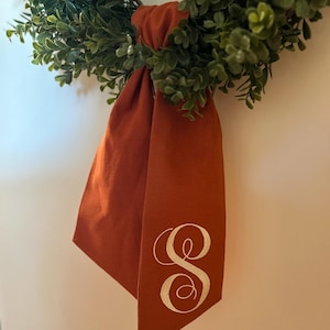 May include: A rust-colored fabric bow with a white embroidered letter "S" hanging on a green wreath.