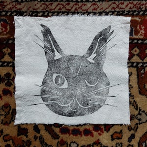 Peut inclure: Illustration en noir et blanc d'un visage de chat imprimé sur un patch de tissu blanc. Le chat cligne de l'œil et a une expression espiègle.
