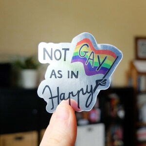 Peut inclure: Un autocollant transparent avec un drapeau arc-en-ciel et le texte "NOT AS IN HAPPY" en lettres noires.