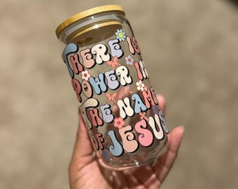 Puede incluir: Un tarro de cristal transparente con tapa de madera. El tarro presenta el texto "THERE IS POWER IN THE NAME OF JESUS" en una fuente retro colorida con detalles florales. El tarro está sostenido en una mano.