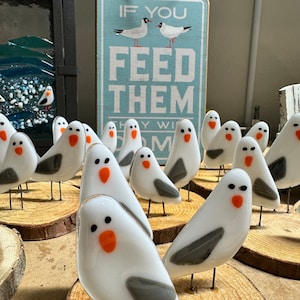 May include: A collection of white glass seagull figurines with gray wings and orange beaks. The figurines are standing on wooden bases. A sign behind the figurines reads "If you feed them they will come".