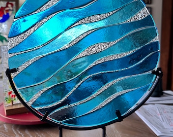Könnte beinhalten: Ein rundes, dekoratives Glasstück mit einem wellenartigen Design in Blau- und Türkistönen, getrennt durch silberne Akzente. Die Glaskunst wird auf einem schwarzen Metallständer präsentiert, der ihre lebendigen Farben und Texturen zur Geltung bringt. Der Durchmesser beträgt etwa 25 cm.