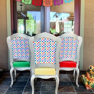 May include: Three silver chairs with colorful polka dot fabric upholstery. The chairs have green, yellow, and red cushions. The chairs are in front of a glass door with a colorful paper garland hanging above.