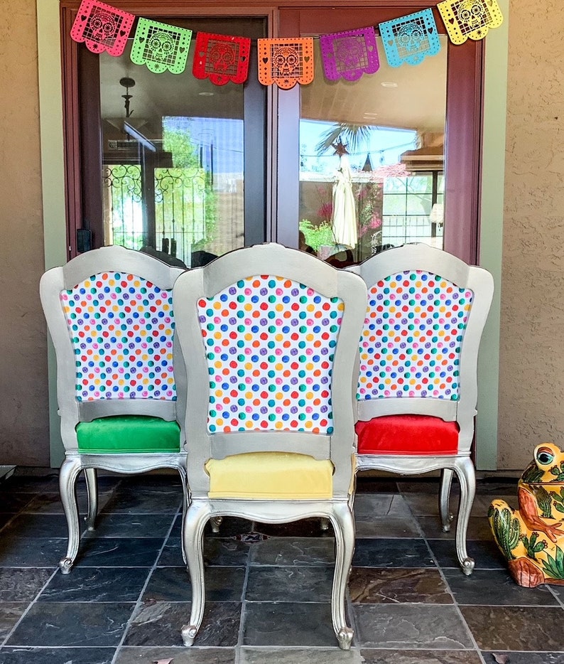 May include: Three silver chairs with colorful polka dot fabric upholstery. The chairs have green, yellow, and red cushions. The chairs are in front of a glass door with a colorful paper garland hanging above.