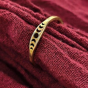 Puede incluir: Un anillo dorado con un diseño de fases lunares en forma de media luna. El anillo se muestra sobre una tela roja oscura, que acentúa los detalles. El diseño del anillo incluye formas de luna llena y creciente.