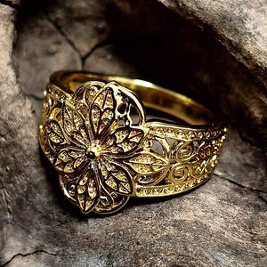 Könnte beinhalten: Ein goldfarbener Ring mit einem kunstvollen Blumendesign. Der Ring hat eine große, detaillierte Blume in der Mitte und aufwendige Filigranmuster am Band. Der Ring wird auf einer strukturierten Holzoberfläche präsentiert.