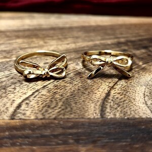 Könnte beinhalten: Zwei goldfarbene Schleifenringe werden auf einer Holzoberfläche präsentiert. Die Ringe haben ein zartes Schleifendesign, wobei die Schleife auf dem Ringband zentriert ist. Der Hintergrund ist eine verschwommene bordeauxrote Farbe.
