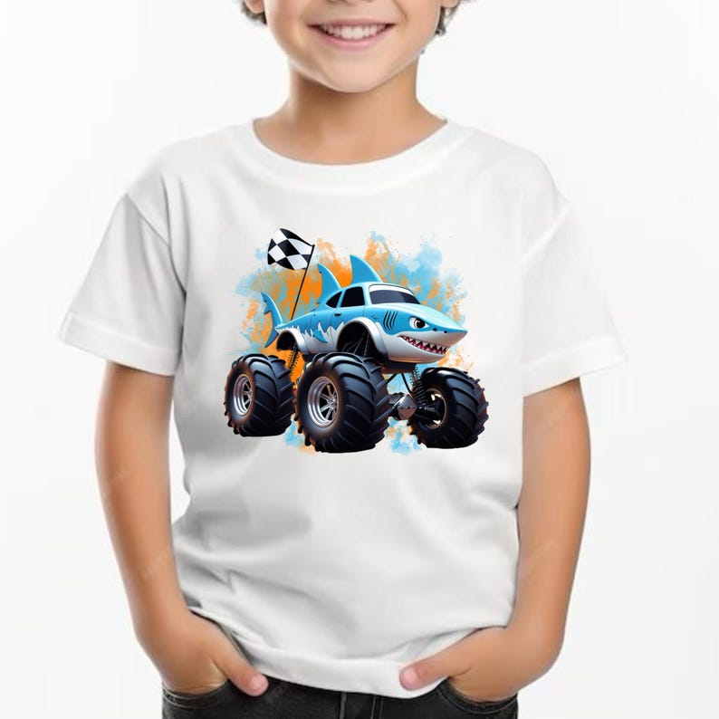 Peut inclure: T-shirt blanc avec un camion monstre de dessin anim&eacute; bleu et blanc avec des dents de requin et un drapeau &agrave; damier. Le camion a de gros pneus et roule sur un fond blanc et orange.