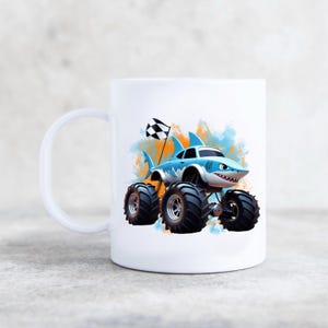 Peut inclure: Une tasse en c&eacute;ramique blanche avec un camion monstre de dessin anim&eacute; bleu et blanc avec des nageoires de requin et un drapeau &agrave; damier. Le camion est sur un fond aquarelle avec des &eacute;claboussures orange et bleues.