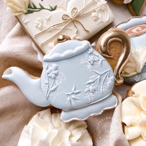 May include: A light blue teapot cookie decorated with white floral details and a gold handle. The cookie is surrounded by other cookies, including a white cookie with gold accents and a brown cookie with a gold handle.