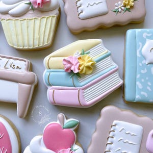 May include: Assortment of decorated sugar cookies in a light color palette. The cookies include a stack of books, a cupcake, a notebook, and an apple. The book cookies are pastel pink, blue, and yellow, with gold accents and floral decorations.