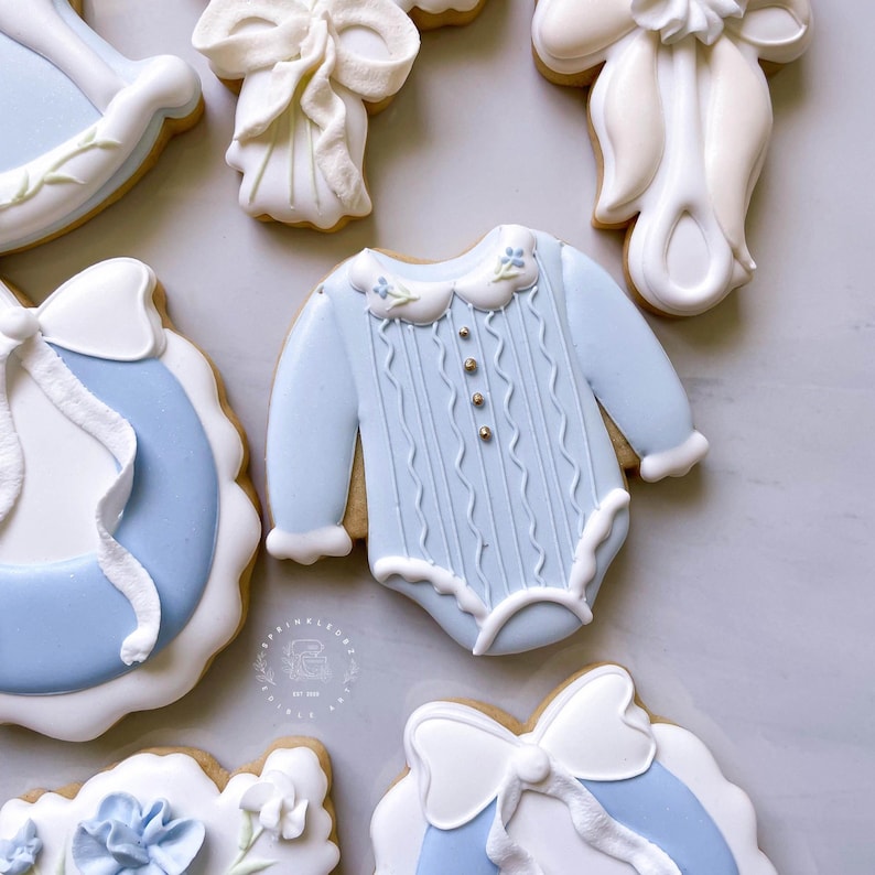 K&ouml;nnte beinhalten: Eine Auswahl an Baby-Themen-Keksen, darunter ein hellblauer Strampler mit goldenen Akzenten, ein Schaukelpferd und Schleifen-Kekse. Verziert mit wei&szlig;em Zuckerguss und blauen Details, ideal f&uuml;r eine Babyparty oder Taufe.