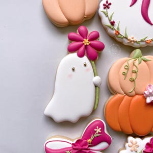 May include: Assortment of decorated sugar cookies. A white ghost cookie holds a pink flower. Other cookies include pumpkins, a floral design, and a pink bow. The cookies are decorated with pink, orange, green, and white icing.